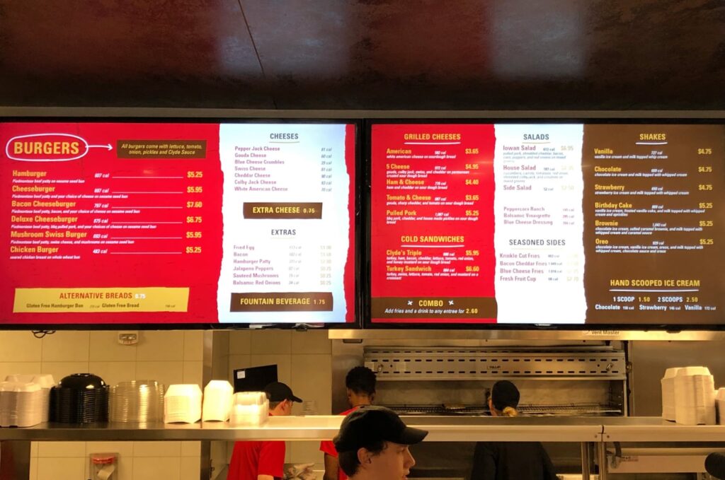 Menu designed for Clyde's Gastro Diner displayed on flatscreen TVs over the counter, featuring burgers, grilled cheeses, salads, shakes, and more.