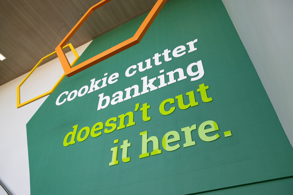 Detail of the Farmer's State Bank stairwell tagline installation, featuring the phrase, "Cookie cutter banking doesn't cut it here" in 3D acrylic lettering. Branded hexagon shapes dangle from the ceiling, tastefully overlapping the message and green wall decal.