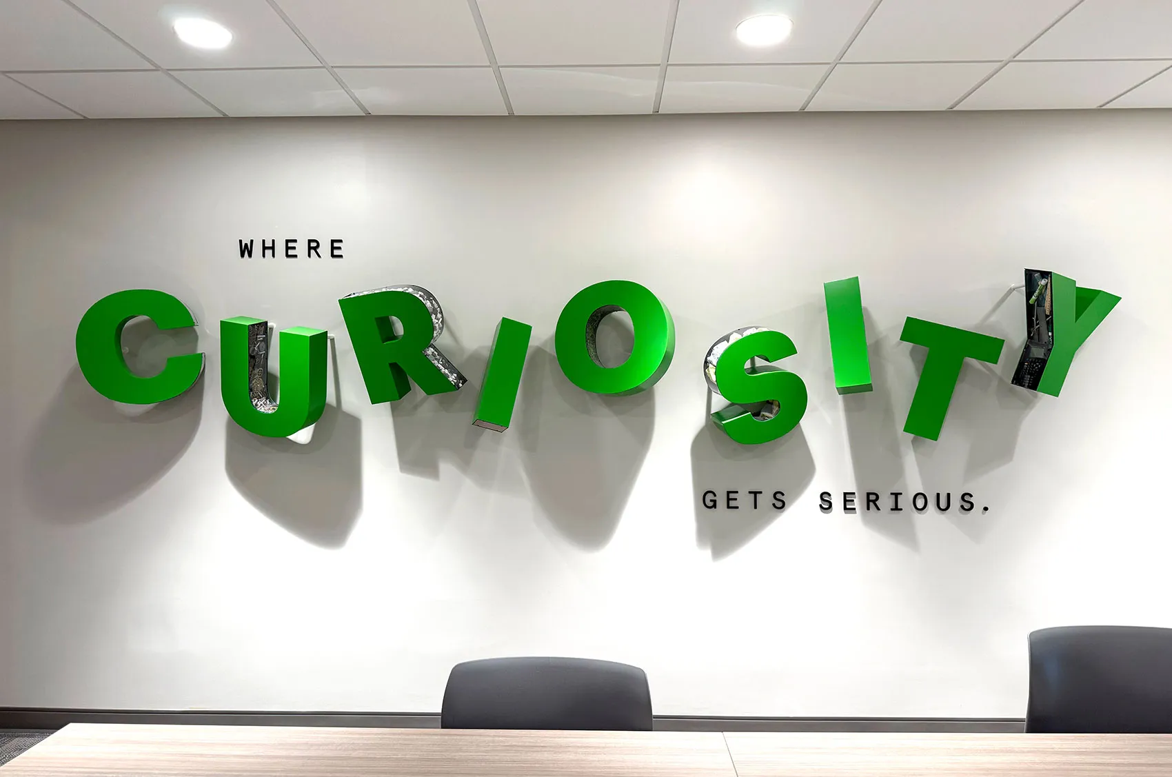 3D green letters spell CURIOSITY on a wall with Where Curiosity Gets Serious slogan. Modern office decor.