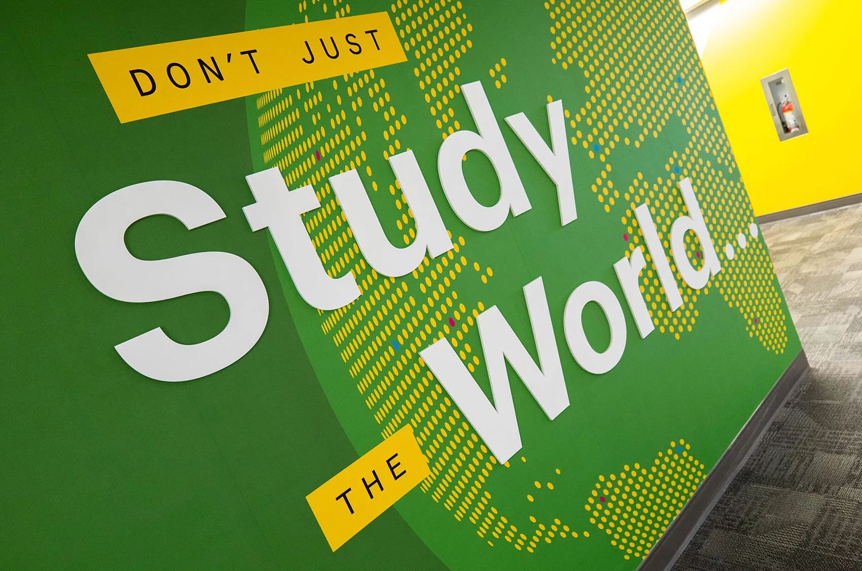 Green wall with Don't Just Study the World text and dotted world map design in yellow and white.